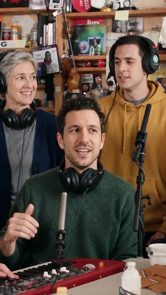 A bookshelf office: a man in a green sweater at a red keyboard sings into a microphone, a silver-haired woman and a young man in a mustard hoodie behind him, all in headphones