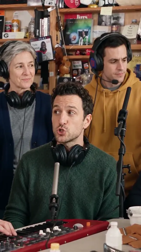The keyboard player turns to the backing singers mid-line while the young man in the mustard hoodie sings