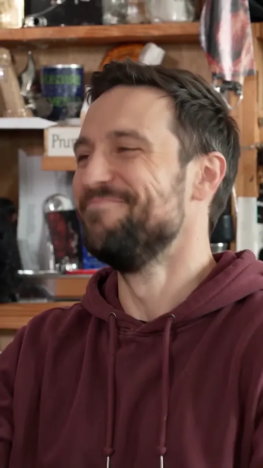 Close-up of a bearded man in a maroon hoodie, eyes squeezed shut, starting to laugh in front of shelves of mugs and records
